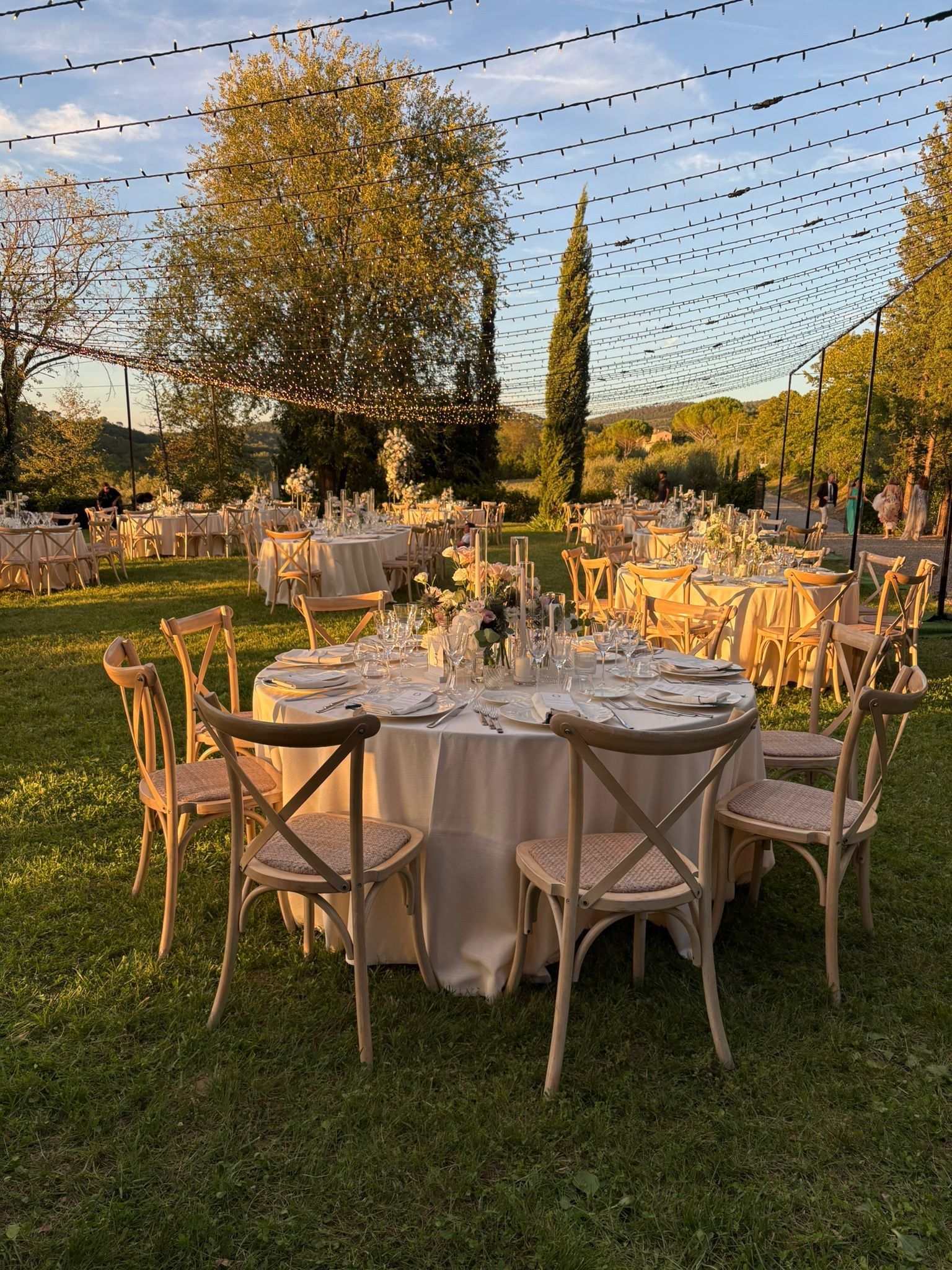 Tavola rotonda apparecchiata sul prato del giardino al tramonto, tra cipressi e file di luci